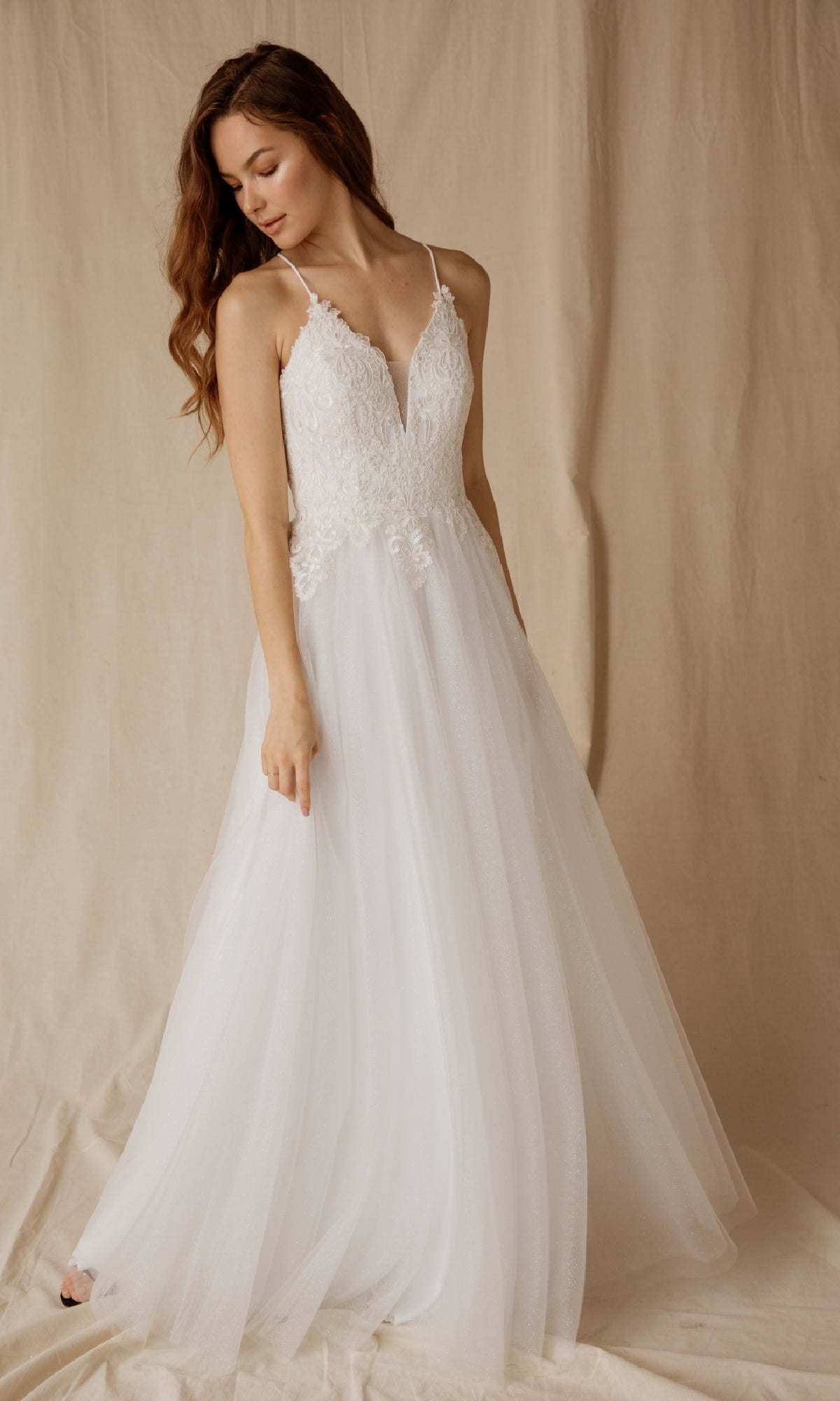 A woman in the Eureka 9901 Lace-Up Long Glitter Prom Ball Gown stands against a beige fabric backdrop, looking down as she gently holds her skirt.